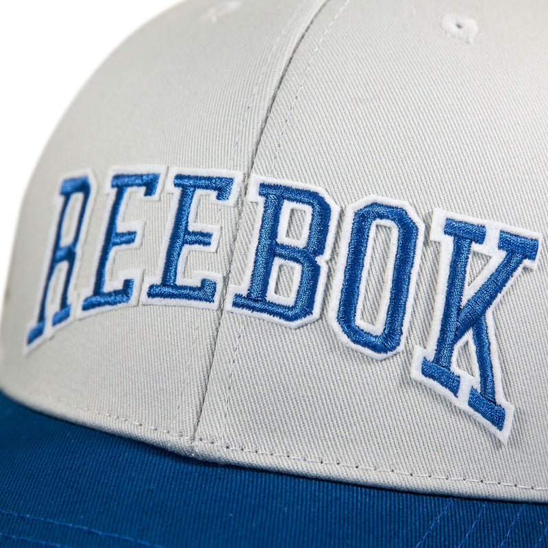 A close-up of the Reebok Gameday Cap, a vintage-inspired cream and blue baseball hat featuring "REEBOK" embroidered in bold blue and white letters on the front.