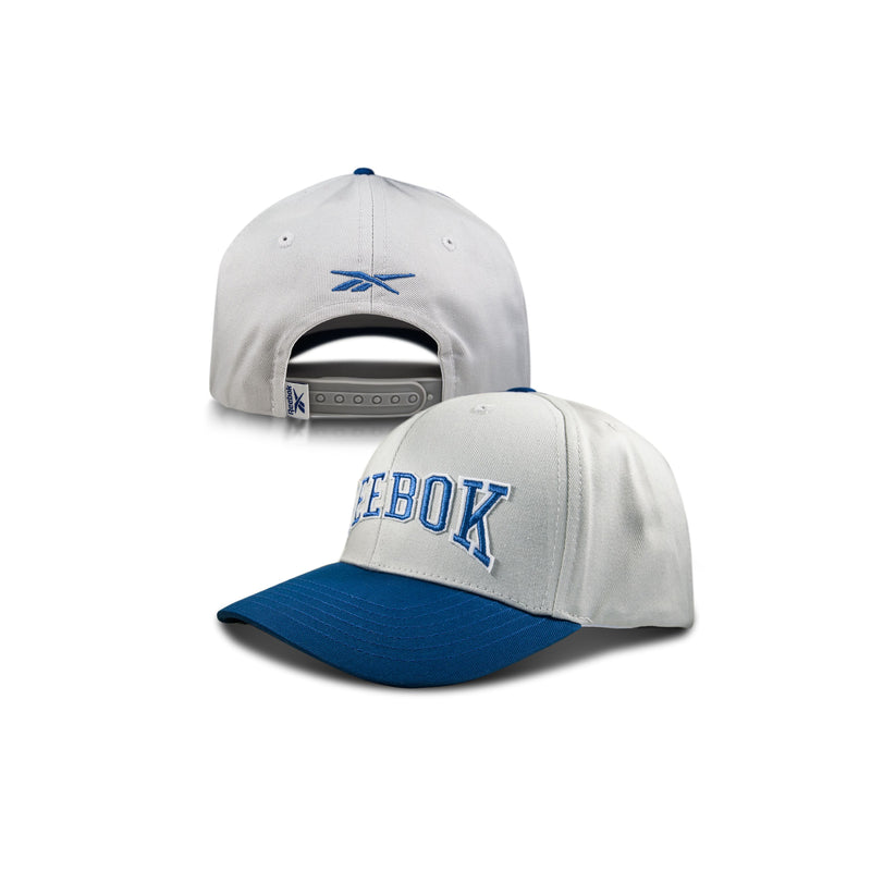 A white and blue Reebok Gameday Cap is shown front and back, featuring bold blue REEBOK lettering on the front, a snapback closure, and an embroidered logo for a vintage-inspired look.