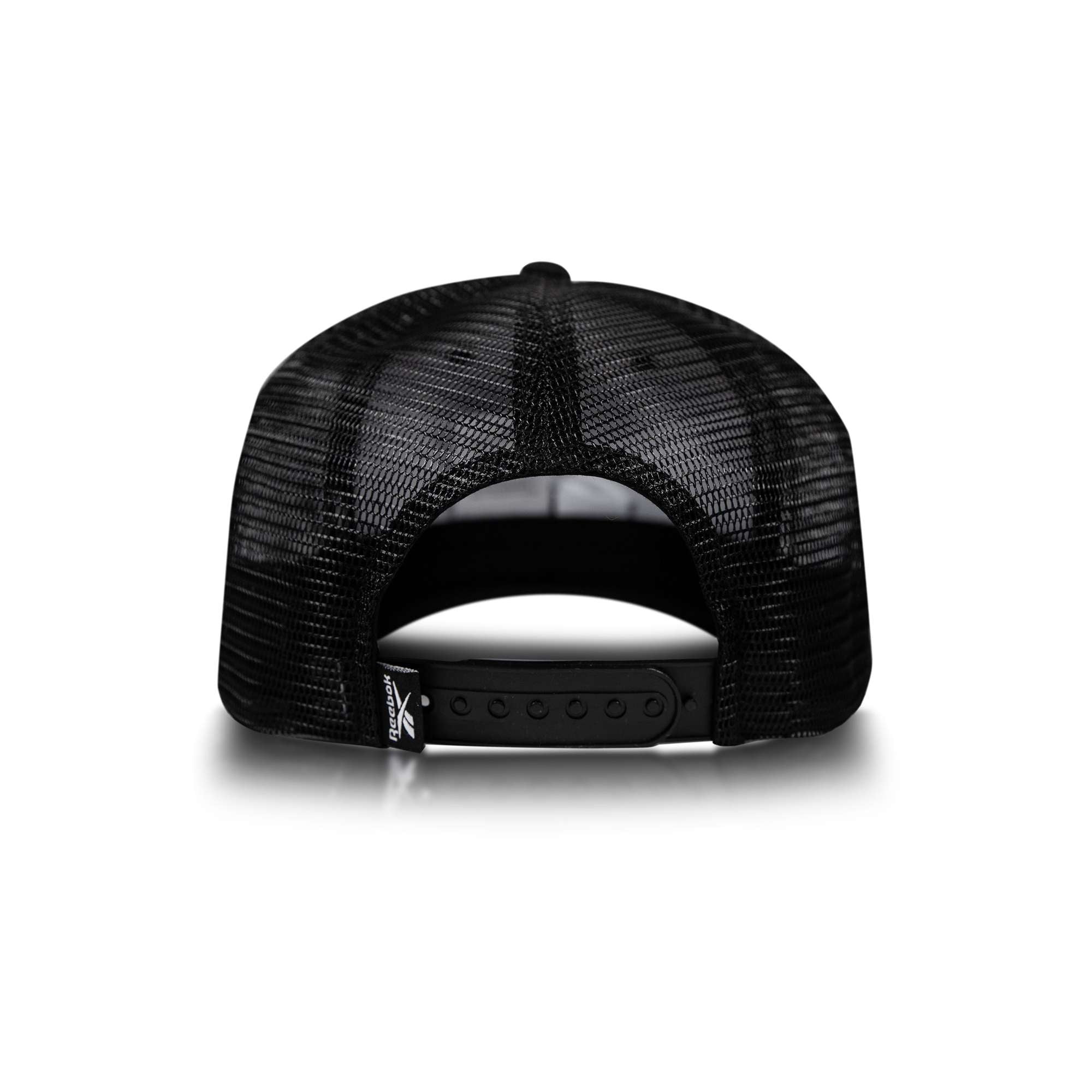 The Reebok Embroidered Vector Trucker Cap is shown from the back, featuring black mesh, an adjustable snapback strap, and a small fabric tag on the lower left of the opening against a white background.