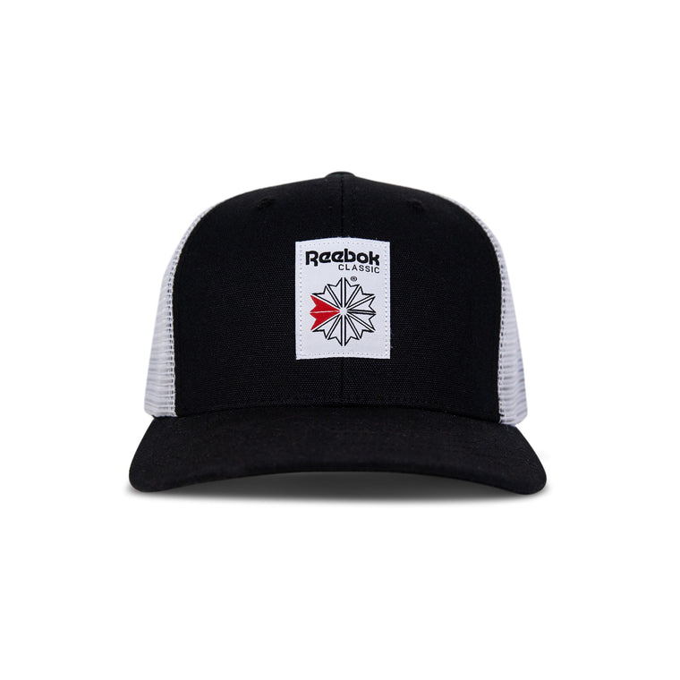 The Reebok Woven Patch Vector Trucker Cap features a black and white design, curved brim, mesh back, adjustable snapback, and a front woven patch with the Reebok logo plus a bold red accent.