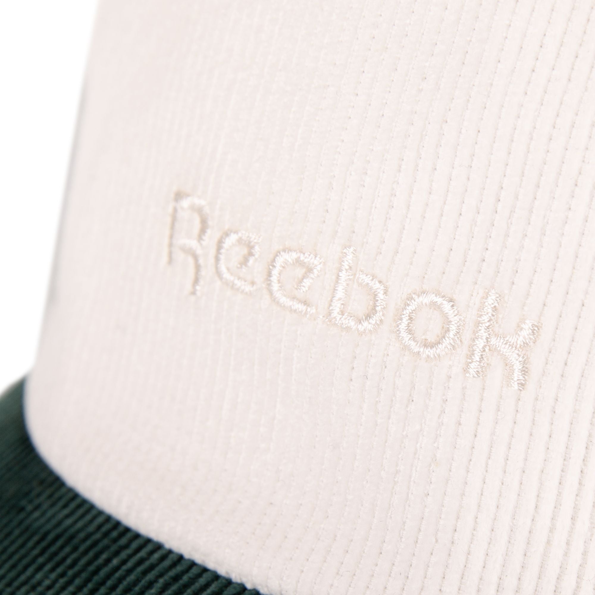 Close-up of white fabric with Reebok’s logo embroidered in white thread. The bottom edge shows a dark green or black corduroy texture, showcasing the classic style of this adjustable Reebok Logo Corduroy Cap.