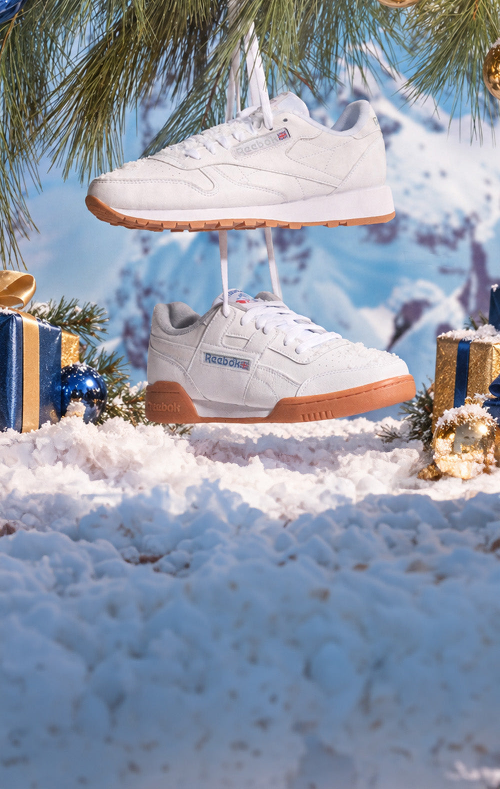 A pair of white Reebok sneakers with gum soles, suspended in a snowy winter scene with pine branches, wrapped gifts, and a blurred mountain background.