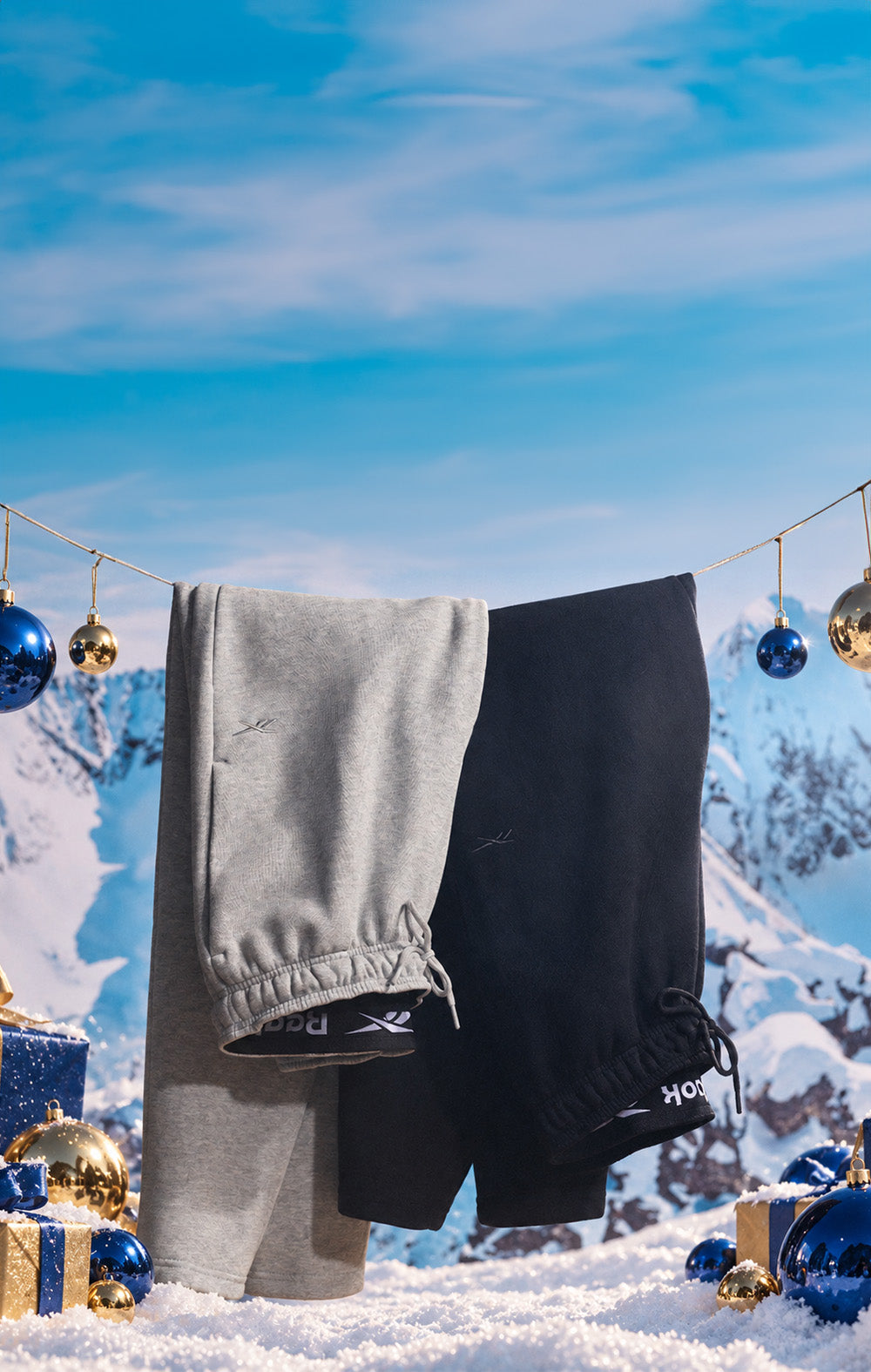 A pair of light gray and black sweatpants hanging on a clothesline outdoors against a snowy mountain backdrop under a blue sky, with Christmas ornaments and wrapped gifts around them.