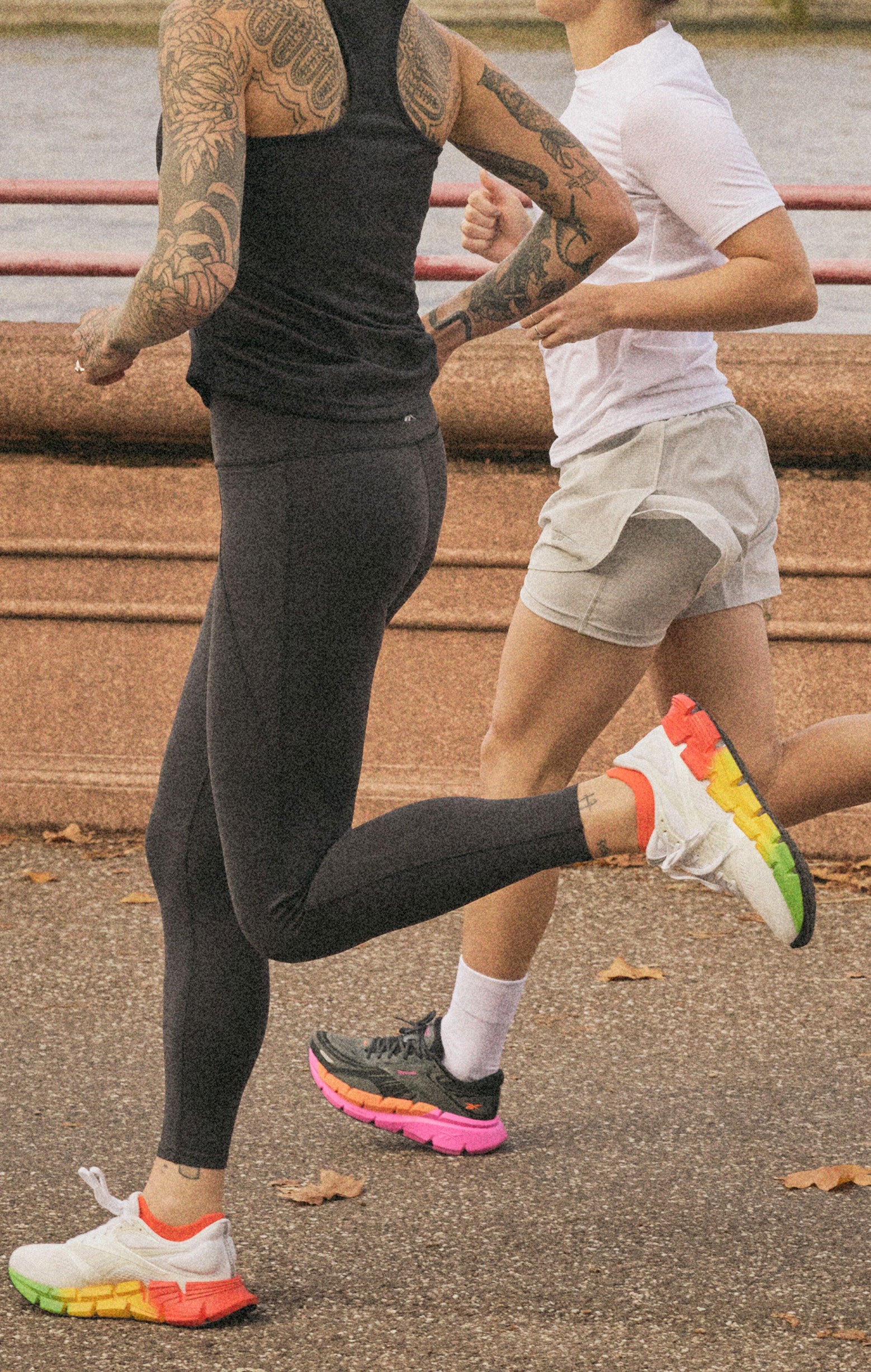 Two people running outdoors on a paved path. One wears black leggings and a black tank top, showing tattoos, and the other wears light shorts and a white t-shirt. Both wear colorful running shoes. A river and railing are in the background.