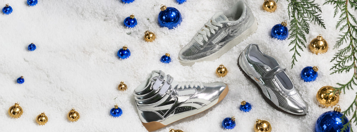 Three silver athletic sneakers arranged on a white snowy surface with blue and gold Christmas ornaments and green pine branches around them.