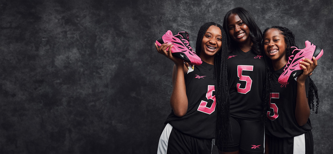 Three girls in black sports jerseys with pink numbers holding pink sneakers against a dark background