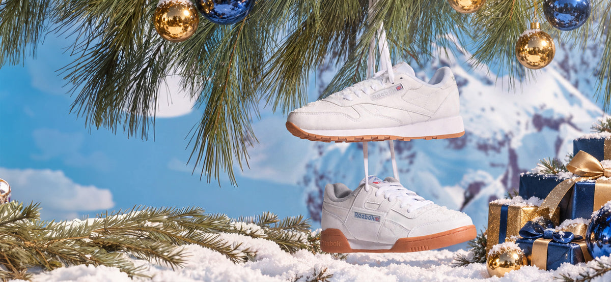 A pair of white Reebok sneakers with gum soles, suspended in a snowy winter scene with pine branches, wrapped gifts, and a blurred mountain background.