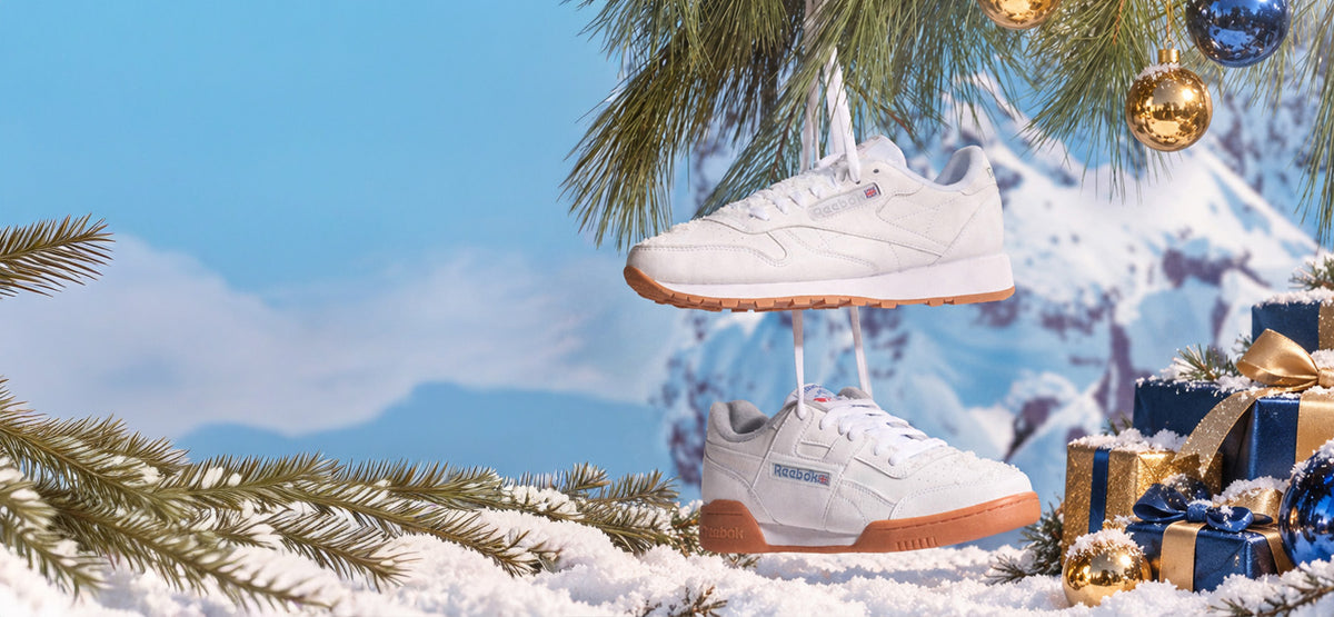 A pair of white Reebok sneakers with gum soles, suspended in a snowy winter scene with pine branches, wrapped gifts, and a blurred mountain background.