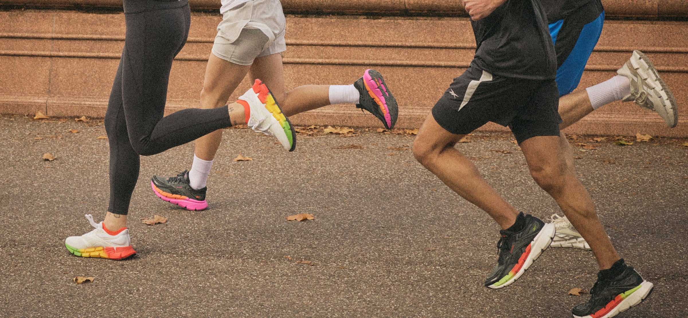 Three people are running outdoors on a paved path, wearing colorful athletic shoes. Only their legs and part of their torsos are visible, with a stone wall and scattered autumn leaves in the background.