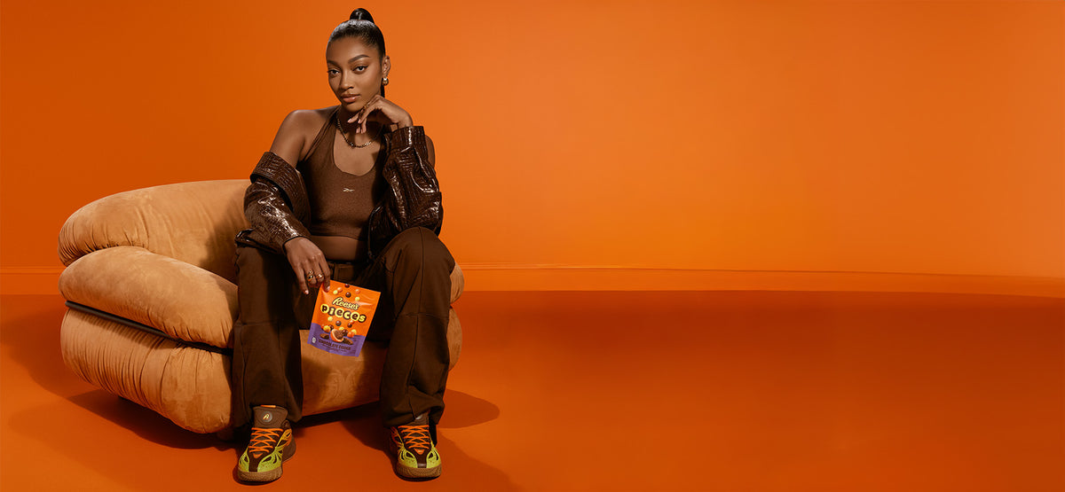 A model sits on a beige armchair against a solid orange studio background, wearing a brown tank top, dark brown pants, a dark patterned jacket, and yellow sneakers, while holding a bag of reeses pieces 