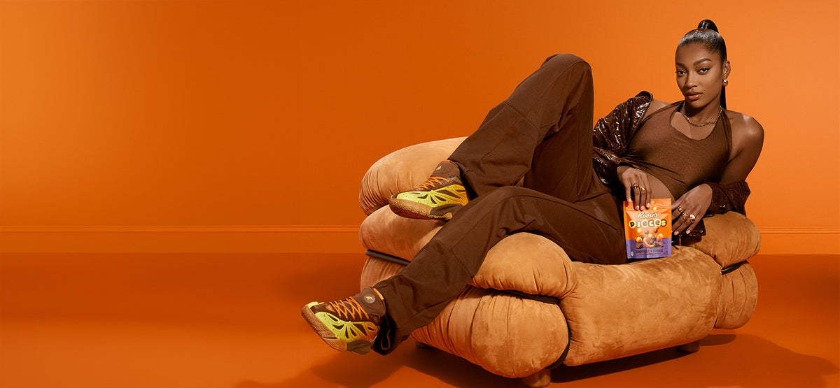 A model reclining on a chunky tan armchair against a solid orange studio background, wearing brown pants and a brown top with yellow sneakers, holding a box of Oreo.