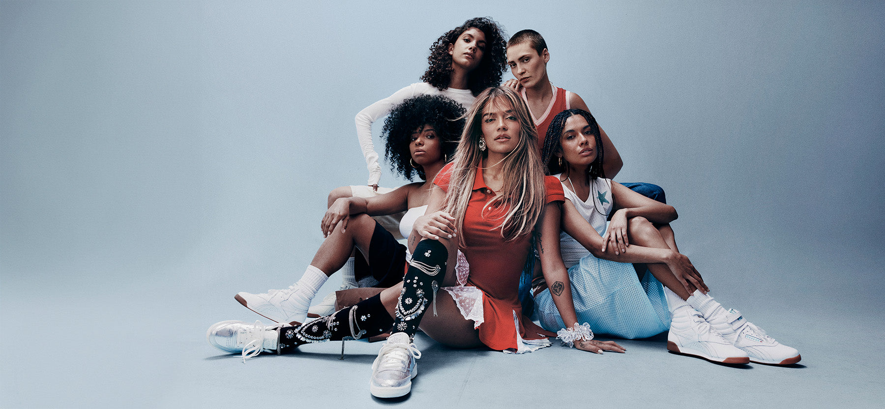 Five fashion models pose together in a studio setting against a cool gray-blue background, wearing colorful outfits including white, red, orange, and light blue, with one model wearing a black-and-white patterned top and white sneakers.