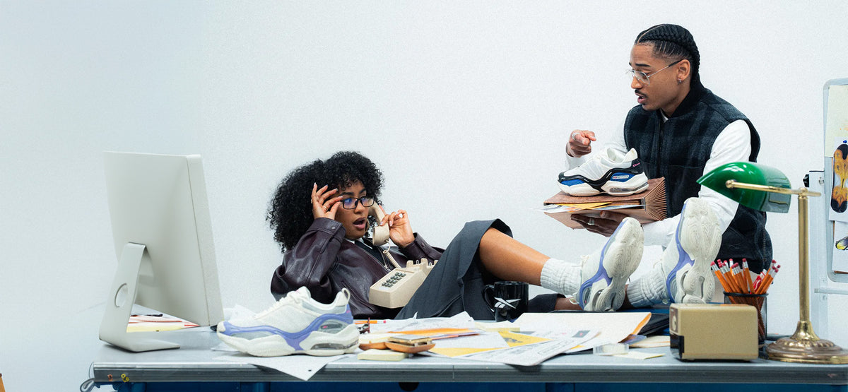 A lifestyle scene showing a man and a woman in a cluttered office setting, both wearing chunky white sneakers with blue and gray accents. The man holds a stack of shoes, and various office items like a computer monitor, lamp, and papers are scattered on the desk.