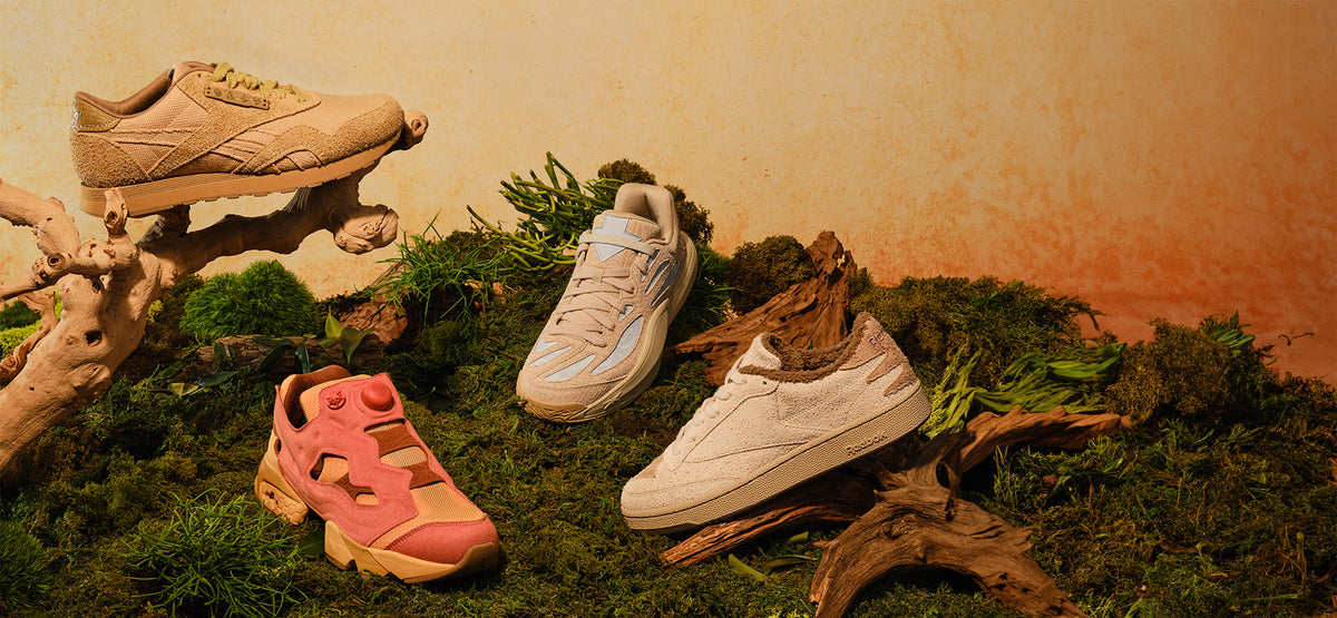 Three pairs of Reebok sneakers displayed in a styled mossy, driftwood setting. The collection includes a beige chunky sneaker on driftwood, a white low-top sneaker on a log, and a pink and beige sneaker resting on green moss.