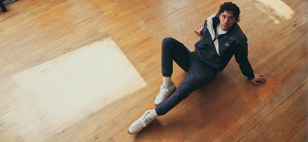 A man wearing a dark navy blue tracksuit with white sneakers sits on a polished wooden floor, looking up towards the camera.