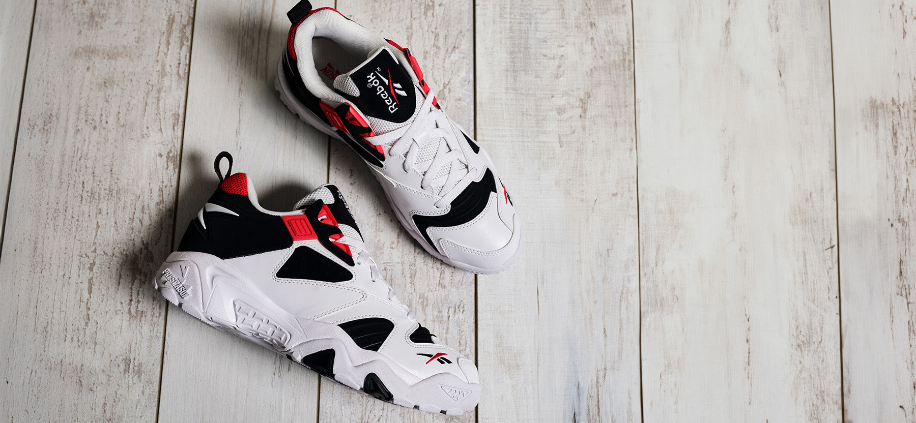 A pair of white, black, and red Reebok sneakers are placed on a white wooden floor, with one shoe upright and the other lying on its side.