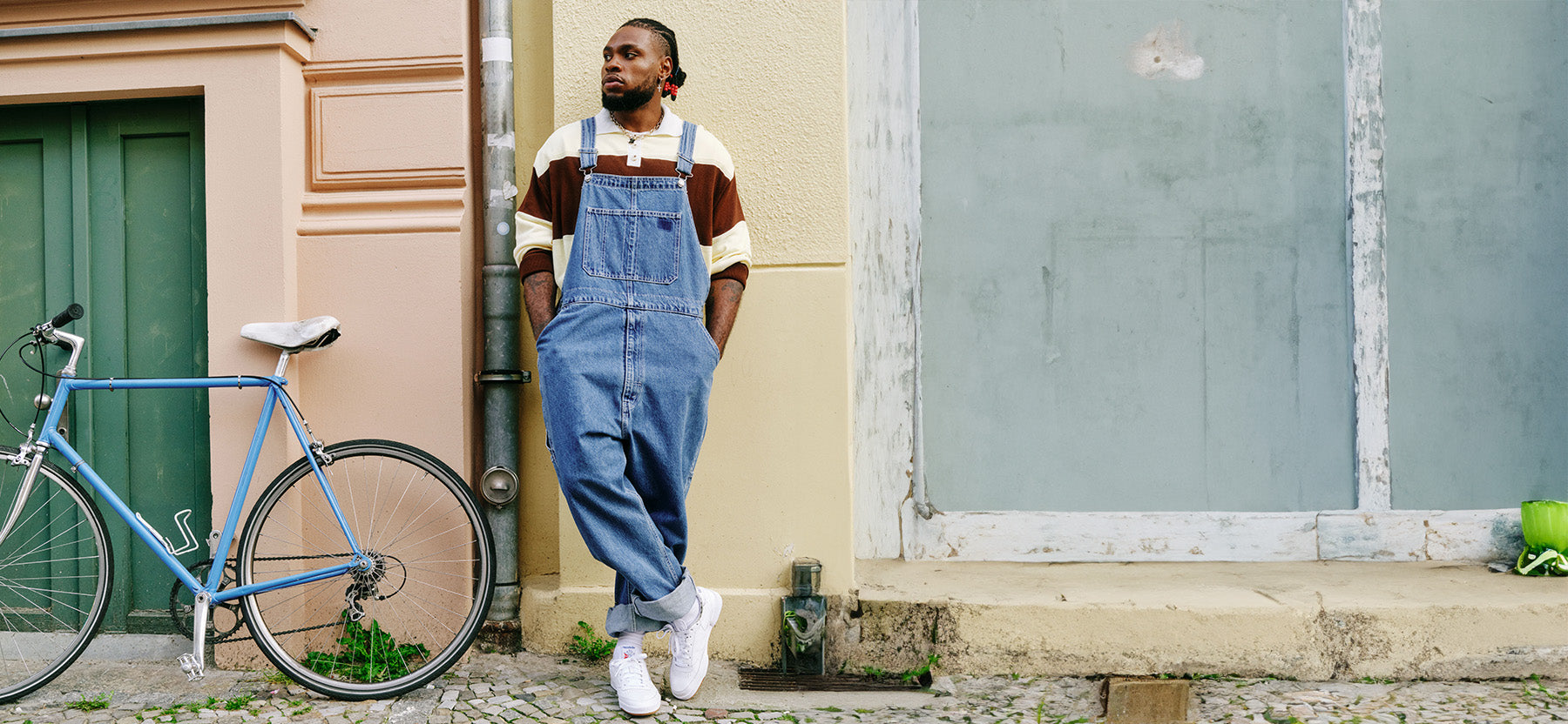 Person wearing blue overalls standing next to a bicycle against a building wall.