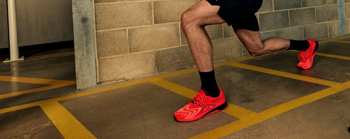 A person in black athletic shorts and black socks runs in a gym wearing bright neon red running shoes, captured from the waist down against a concrete block wall and yellow painted floor markings.