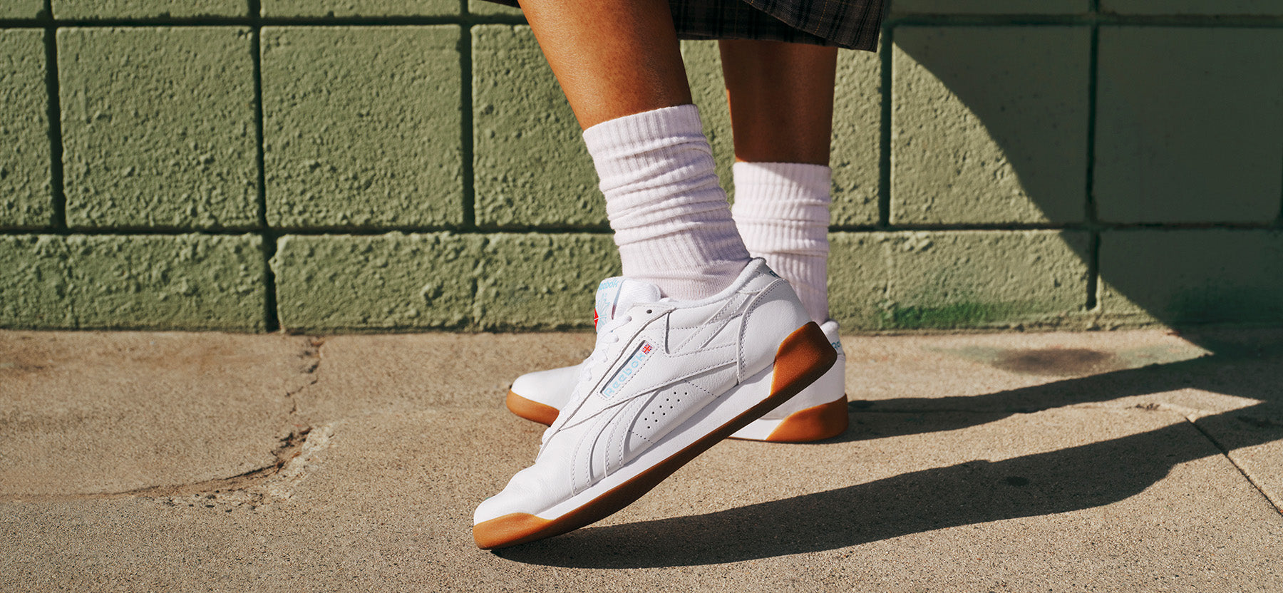 White sneakers with brown soles worn by a person against a green brick wall.