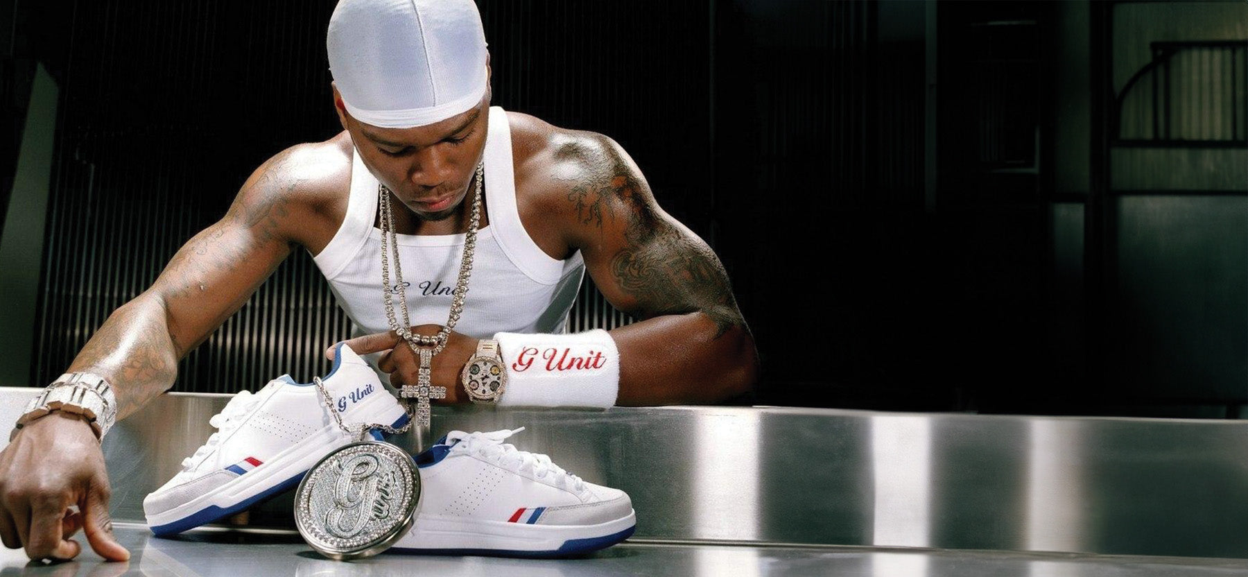 A man in a white cap and white tank top leans over a metallic surface with a pair of white sneakers and a large coin. The sneakers have red and blue accents and the cuff is printed with 'G-Unit' in red.