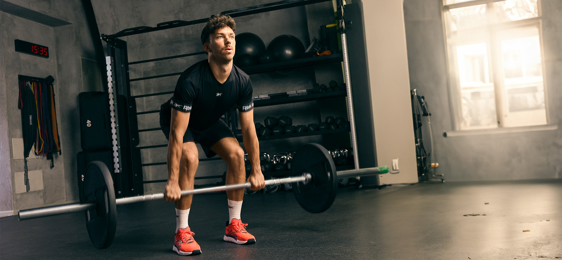 A man in a gym wearing a black athletic t-shirt and shorts, lifting a barbell while wearing bright orange-red running shoes. The background shows a weight rack, gym equipment, and a window.