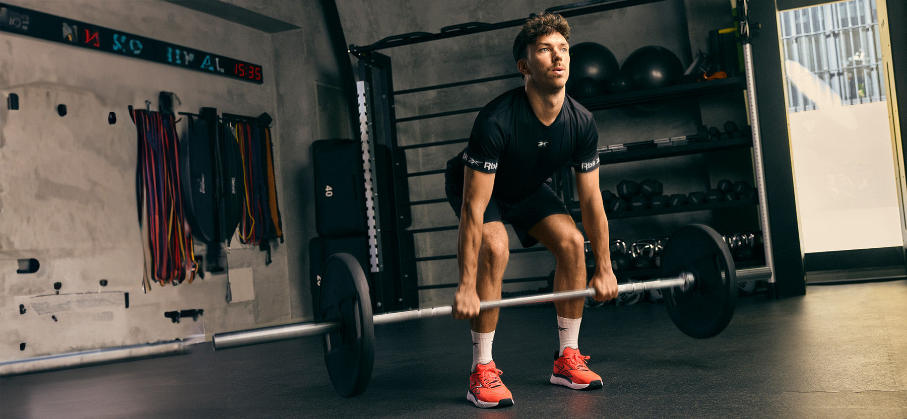 A man in a gym wearing a black Nike athletic shirt and shorts, lifting a barbell. The Nike logo is visible on his shirt.