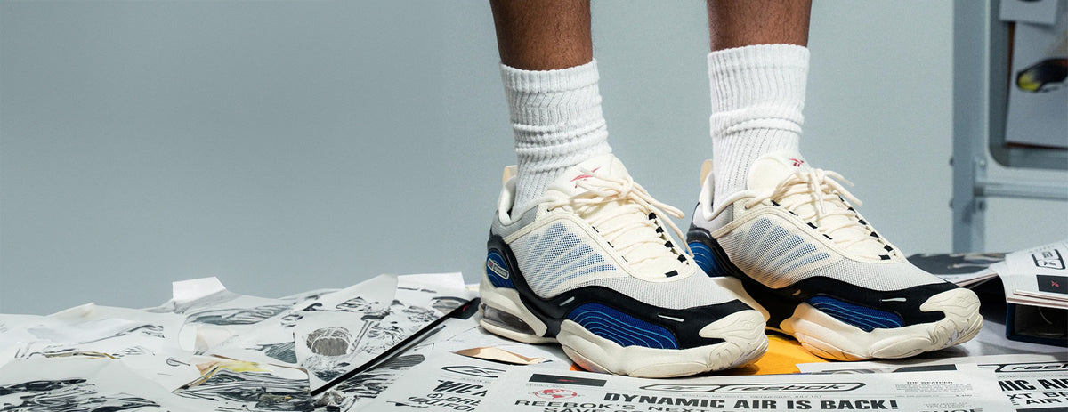 A person wearing a pair of chunky off-white sneakers with blue and black accents, paired with white crew socks, standing on a floor covered in printed papers.