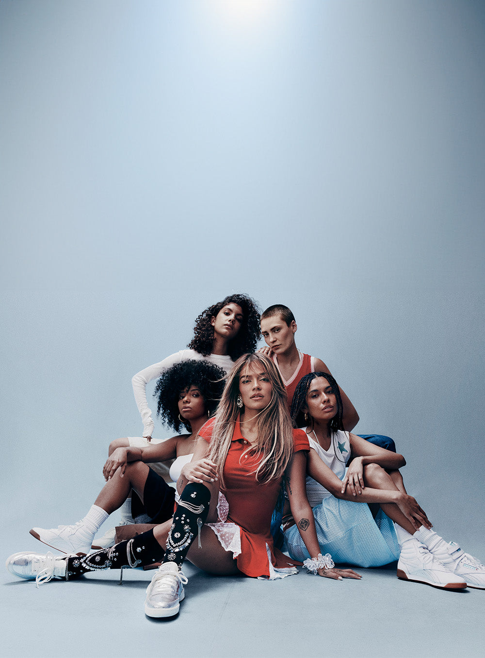 Five models pose together on the floor against a light blue-gray studio background, wearing casual outfits in white, orange, red, and light blue, with one model wearing a black prosthetic leg.