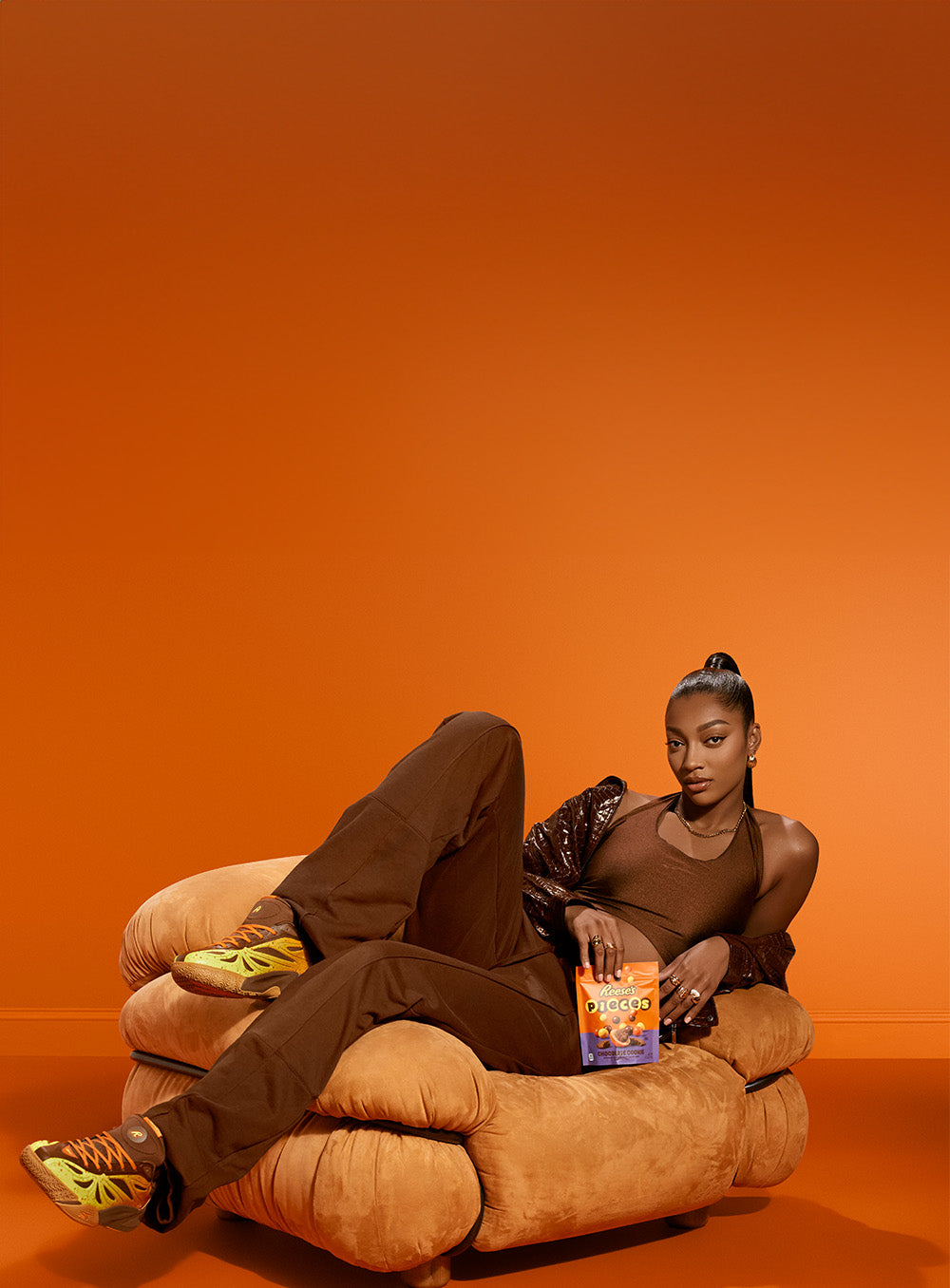 A model reclining on a low, rounded tan chair against a solid orange studio background, wearing a brown tank top, brown pants, and bright yellow-green sneakers, while holding a bag of Frito-Lay chips.