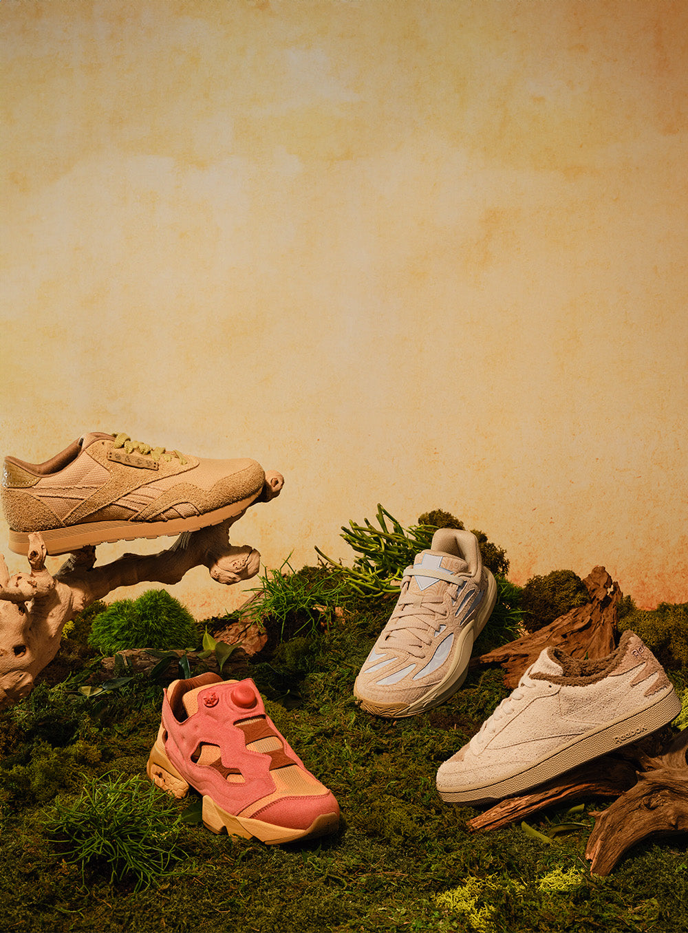 Three Reebok sneakers displayed in a styled natural setting with moss, wood, and a textured beige wall. The shoes include a beige chunky sneaker resting on a rock, a white sneaker on a log, and a pink and beige sneaker in the foreground.