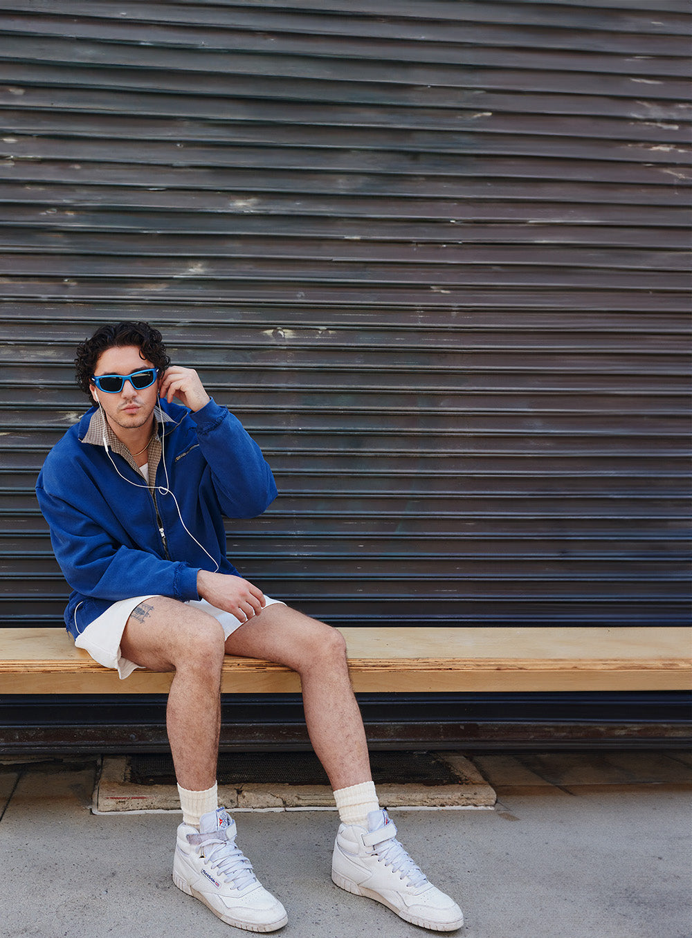 A man wearing a blue zip-up hoodie, white shorts, white sneakers, and sunglasses sits on a wooden bench in front of a dark corrugated metal wall.