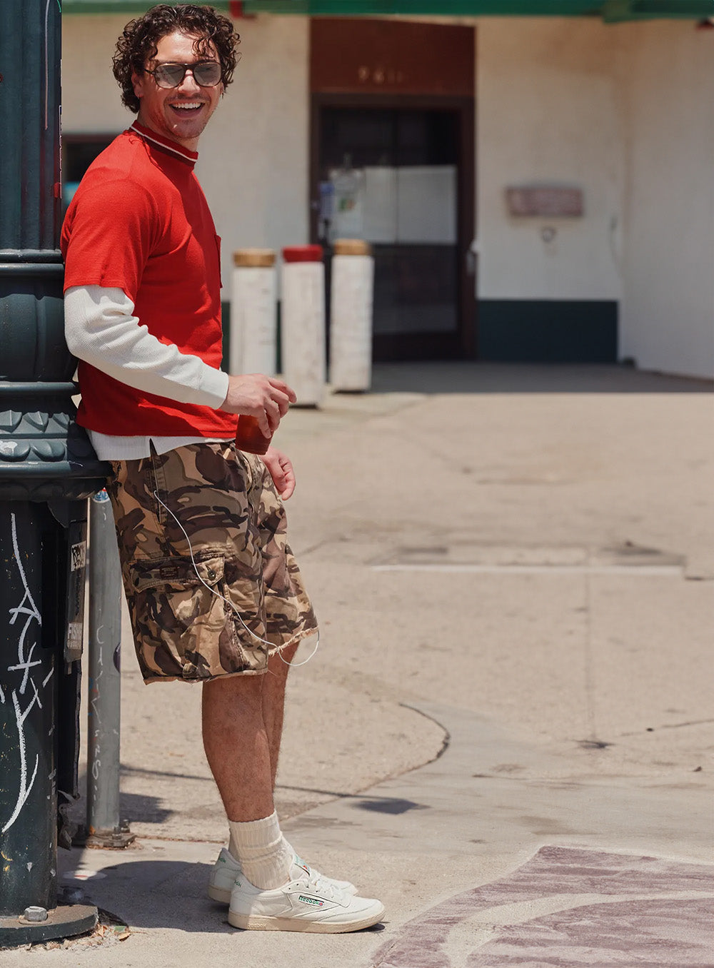 A man wearing a red long-sleeve shirt over a white long-sleeve, camouflage cargo shorts, white socks, and white sneakers, leaning against a dark green pole in an outdoor urban setting.