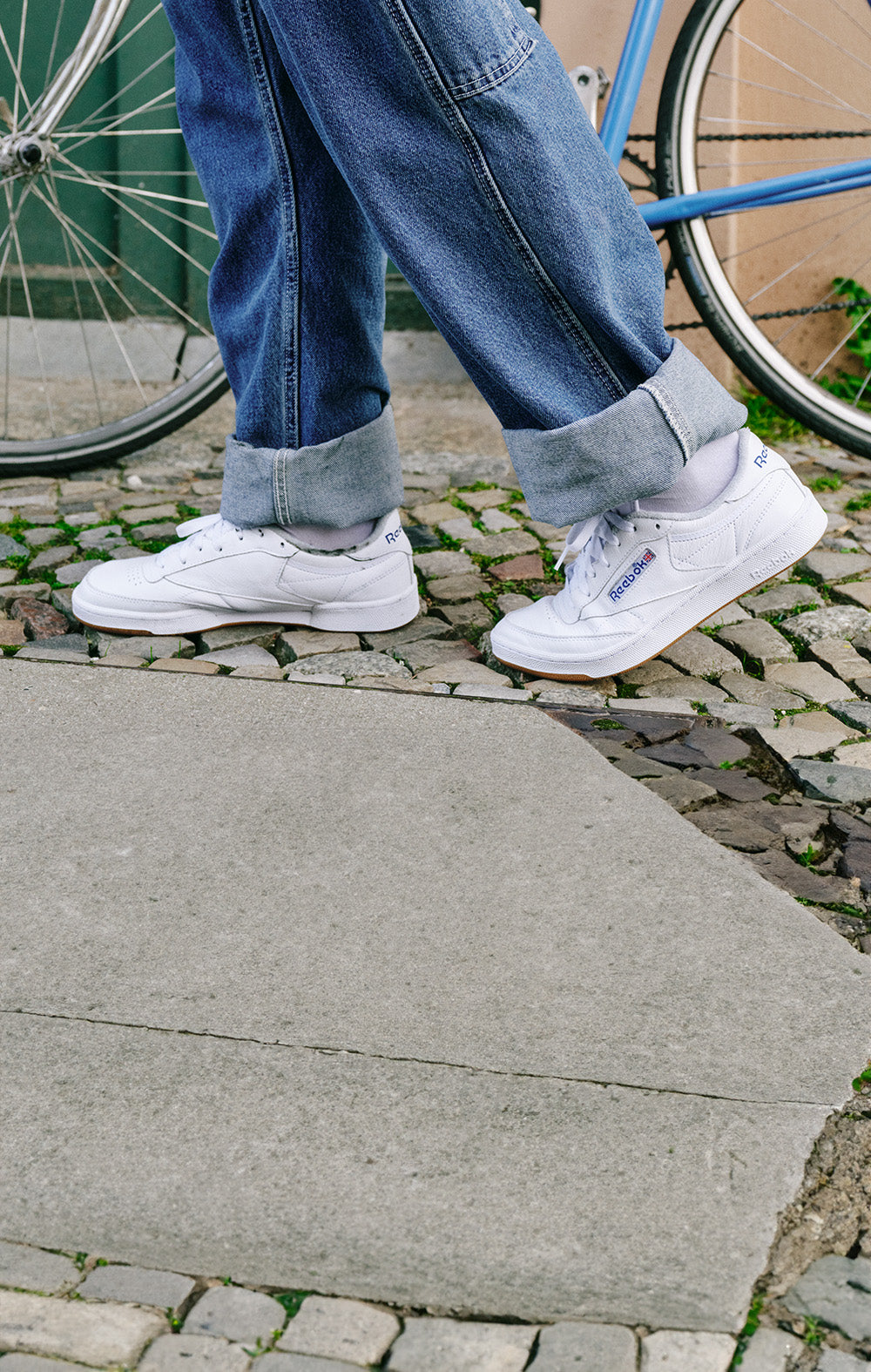 White sneakers worn with blue jeans on a stone pavement, with a bicycle in the background.