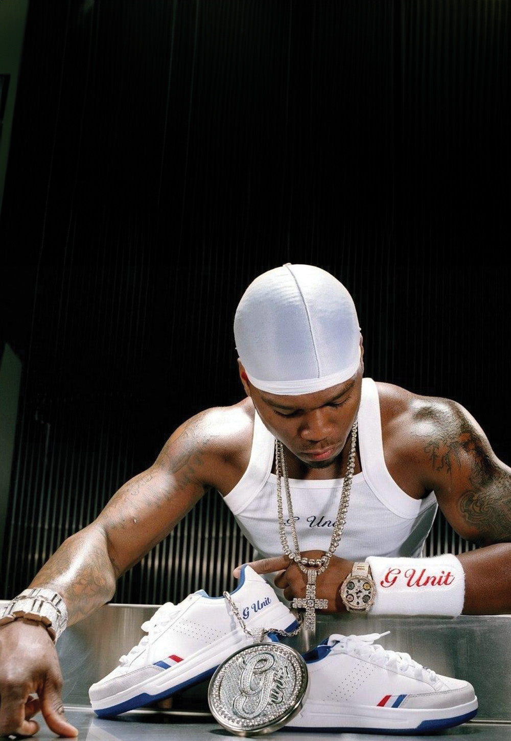 A man wearing a white cap, white tank top, and silver jewelry leans over a pair of white G-Unit sneakers with blue and red accents, with a large silver medallion in the foreground against a dark background.