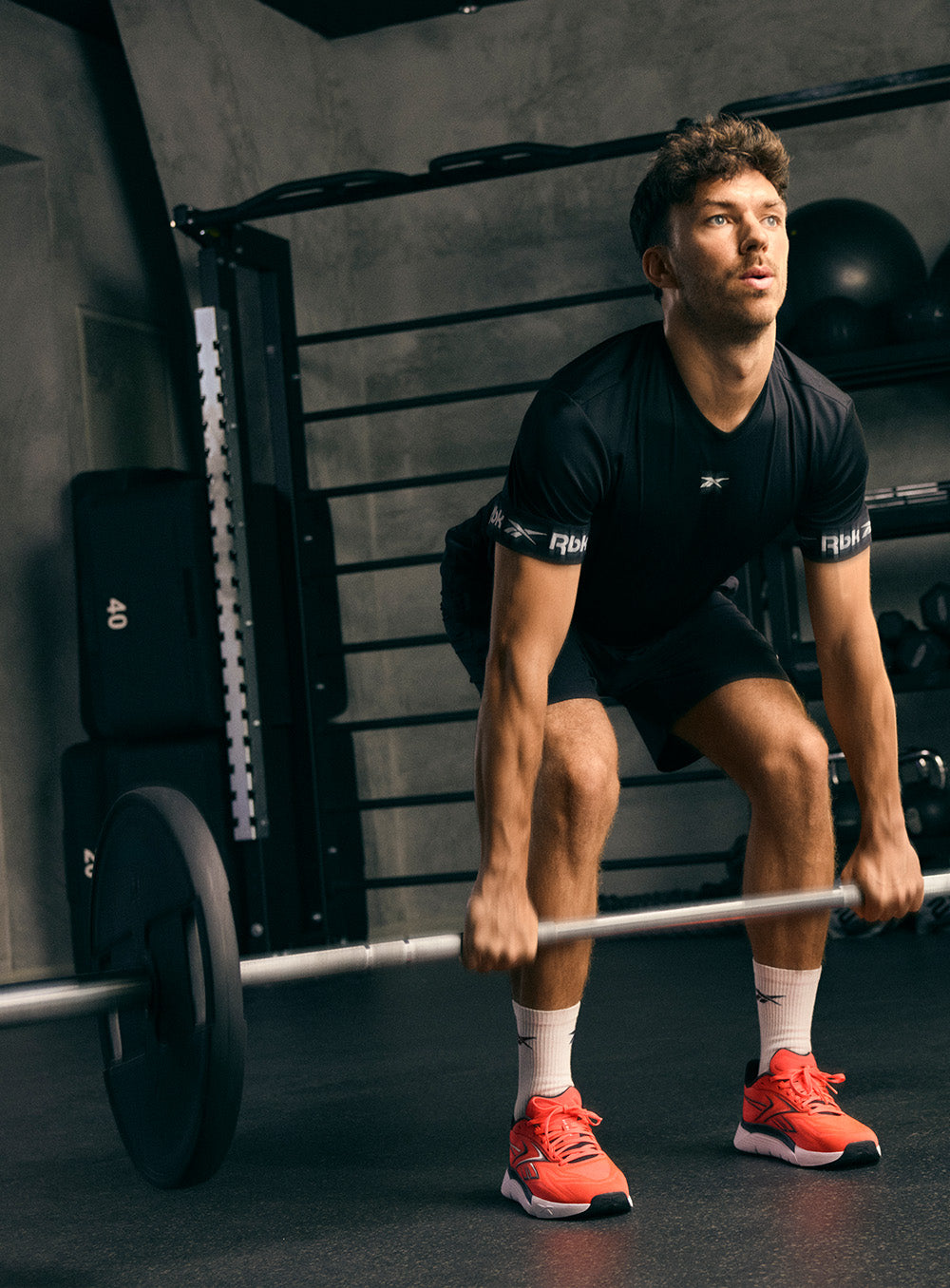 A man in a gym wearing a black athletic t-shirt and shorts, lifting a barbell. The shirt features a small white logo on the chest and sleeve, with 'Fit' printed on the sleeve cuff.