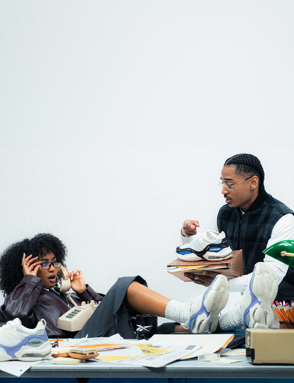 A fashion editorial photo featuring a man and a woman in a stylized, relaxed office setting. The man sits on a desk piled with papers and books, wearing a black vest over a white long-sleeve shirt and white sneakers with blue accents.