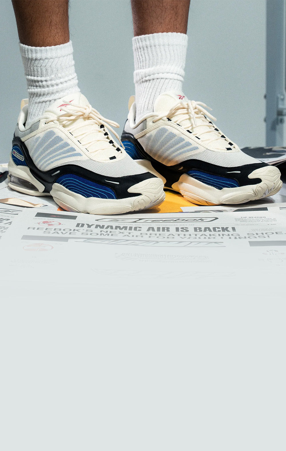 A person wearing a pair of chunky off-white sneakers with blue and black accents, paired with white crew socks, standing on a floor covered in printed papers.
