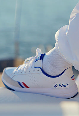 A close-up of a white athletic sneaker with blue and red accents, worn with a white sock and white pants. The shoe has 'E Unit' written in blue script on the side, and the background is a soft, blurred outdoor scene.