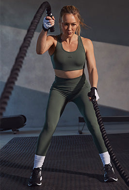 A woman in an olive green athletic set consisting of a sports bra and leggings, holding battle ropes in a gym setting.