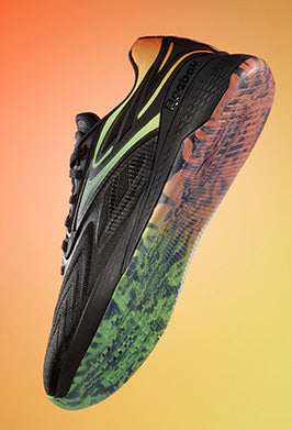 A black athletic shoe with green and yellow accents and a multicolored sole, shown from a low angle against a gradient orange-yellow background.