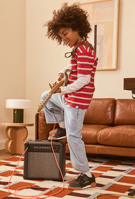 A child with curly hair stands in a living room, playing an electric guitar connected to a black guitar amplifier. The child wears a red and white striped long-sleeve shirt, light blue jeans, and black sneakers.