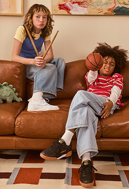 Two children sitting on a brown couch, one holding a broom and the other a basketball, with a dinosaur toy on the floor.
