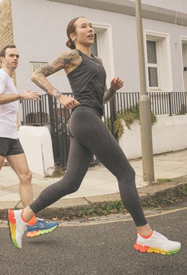 A woman in athletic wear jogs on a city sidewalk, while a man in shorts and a T-shirt runs behind her. They are passing residential buildings and a streetlamp.