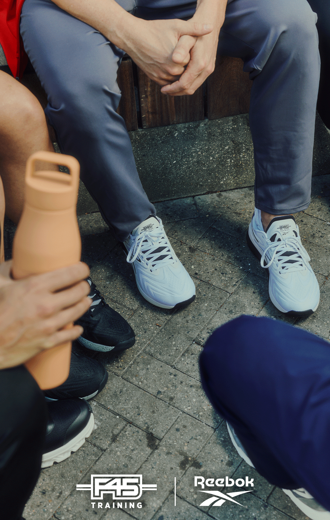 Three people sitting together outdoors, dressed in athletic wear and sneakers. One holds a tan water bottle. The F45 Training and Reebok logos appear at the bottom of the image.