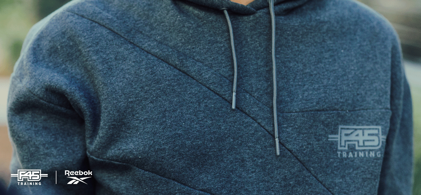 Close-up of a person wearing a blue-gray F45 Training hoodie with diagonal stitching. The F45 Training and Reebok logos are visible in the bottom left and on the chest. The hoodie has drawstrings.