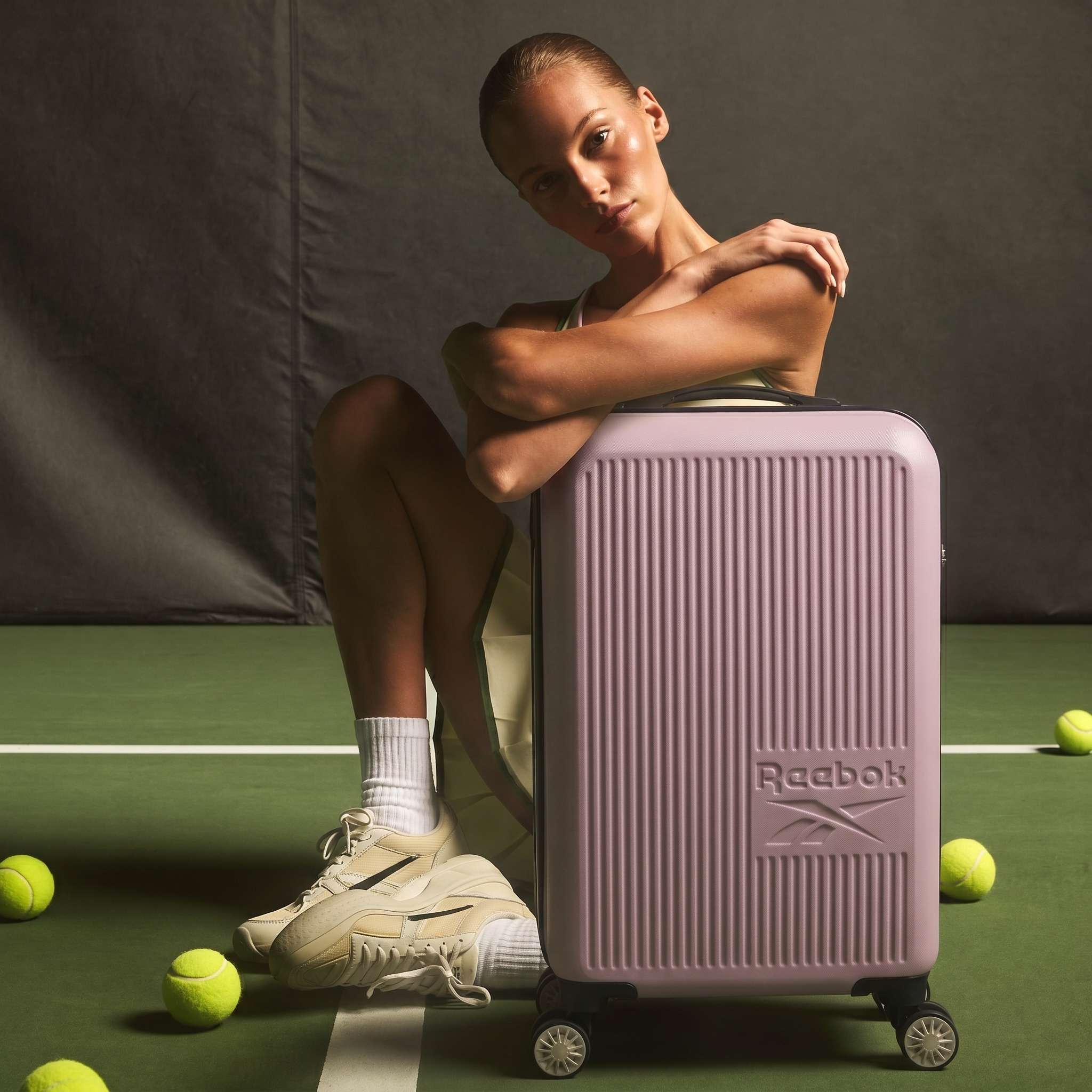 A woman in tennis attire sits on a green court with the pink Reebok Action Duo 2 Piece Luggage Set. Tennis balls and a racquet surround her as she rests her arms on the luggage, wearing white sneakers and gazing confidently at the camera.