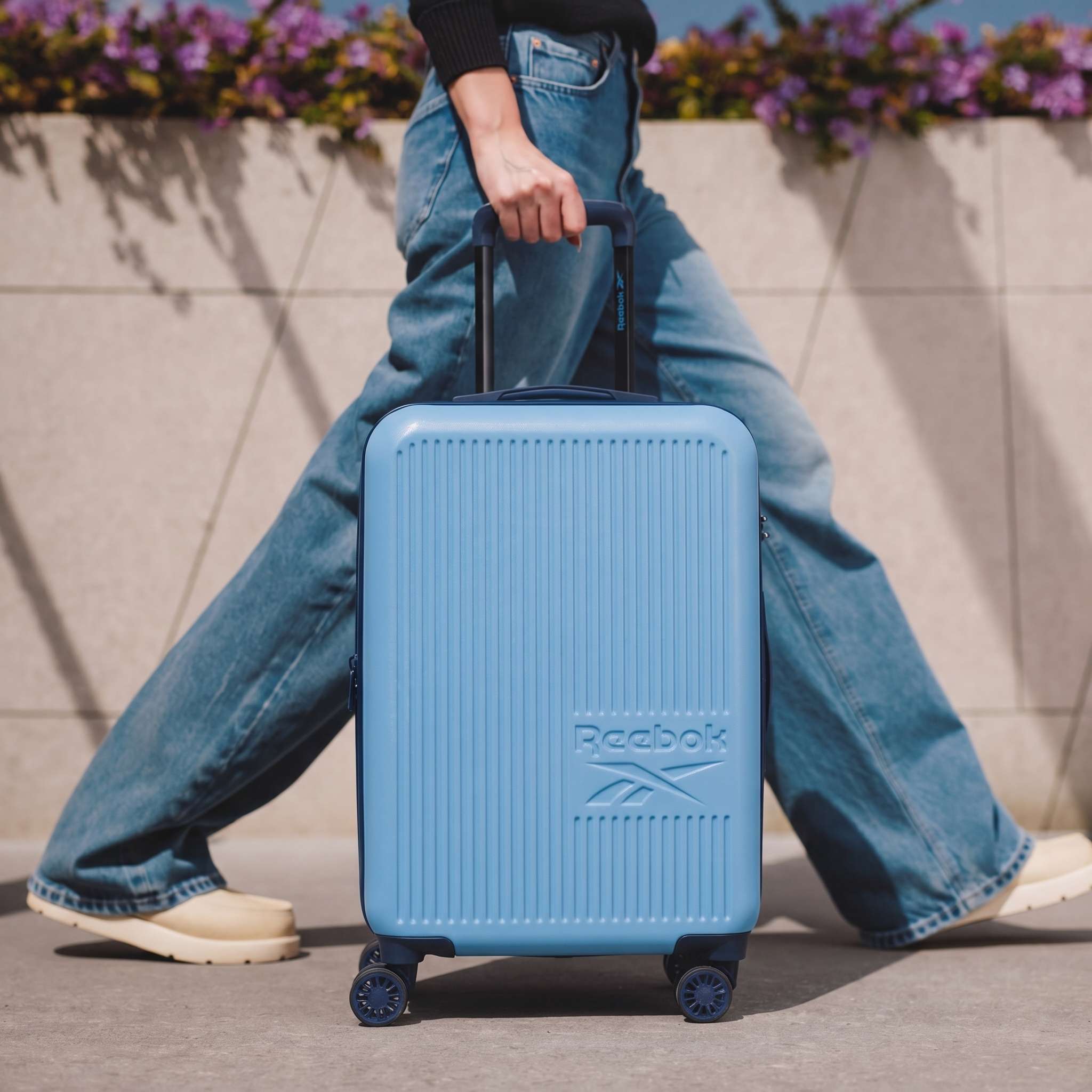 A person in light-colored shoes and blue jeans pulls a light blue Reebok Action Duo 2 Piece Luggage Set with spinner wheels outdoors, with purple flowers in the background.