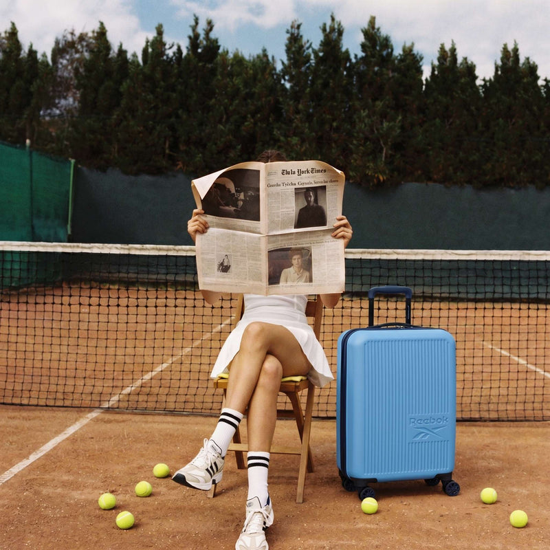 Wearing a white tennis outfit, a person reads a newspaper on a court chair beside scattered tennis balls and the Reebok Action Duo 2 Piece Luggage Set. The net and trees are visible in the background.