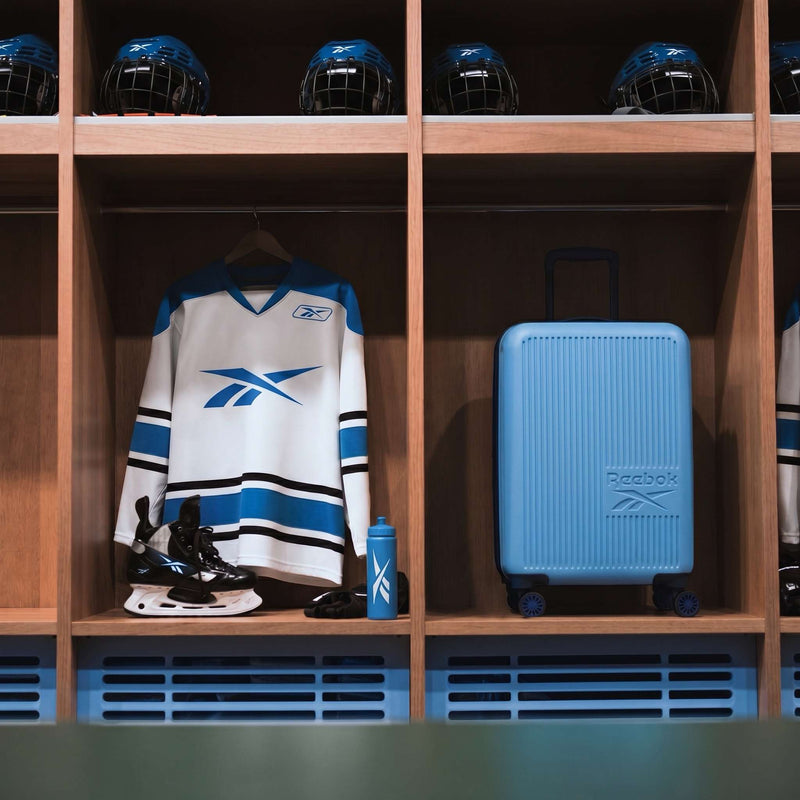 A hockey locker displays a white and blue jersey, black skates, a blue water bottle, several blue helmets on the top shelf, and the Reebok Action Duo 2 Piece Luggage Set with spinner wheels.