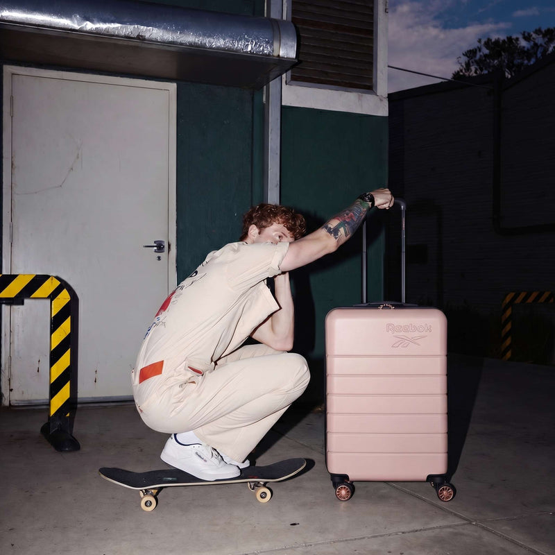 A person in a beige jumpsuit squats on a skateboard at night, holding Reebok's Captain 2 Piece Luggage Set with spinner wheels. Yellow and black safety barriers and a green wall are in the background.